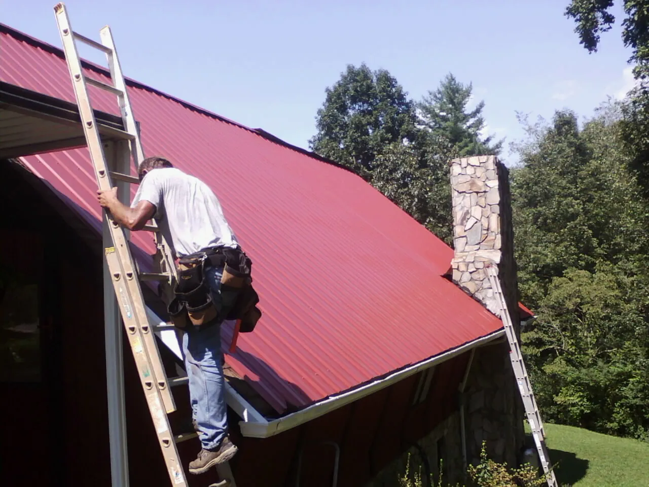 Roofer on a ladder beside a red metal roof and stone chimney — Soffit & Fascia Repair by Roofing Brothers in Brewer